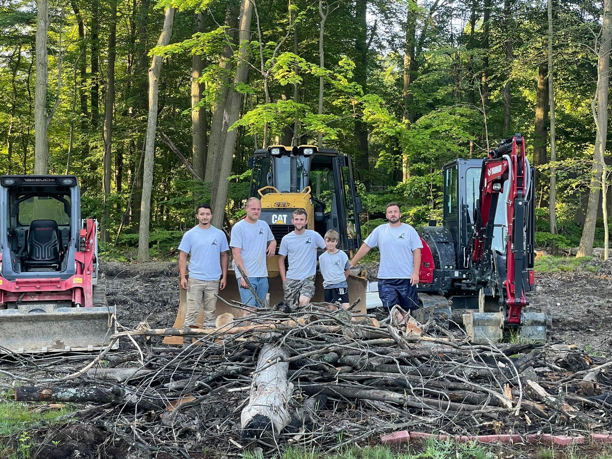 Four adults and one child stand in front of construction equipment and a pile of branches and logs in a wooded outdoor area.