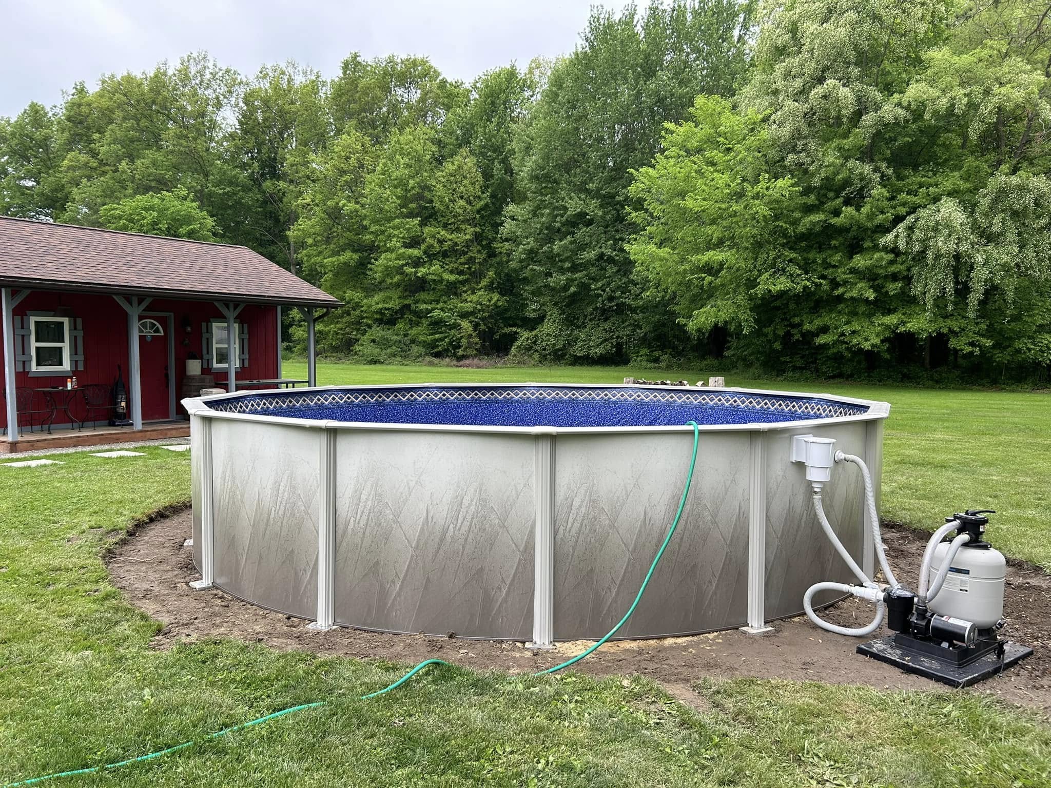 An above-ground swimming pool with a filter system, set up on a grassy lawn near a red shed, surrounded by trees.