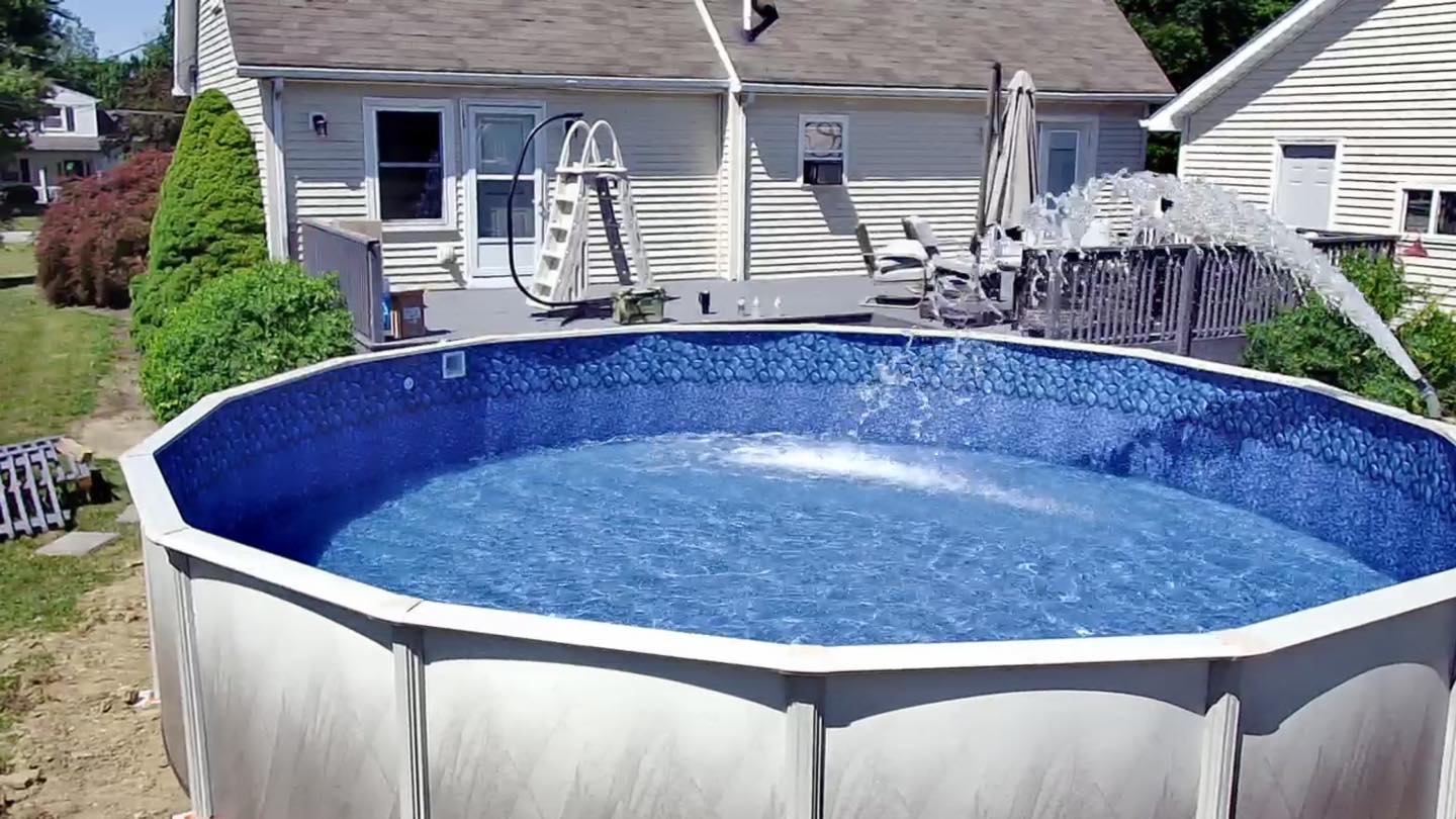 Above-ground swimming pool filled with clear blue water in a backyard, with a house and patio furniture visible in the background.