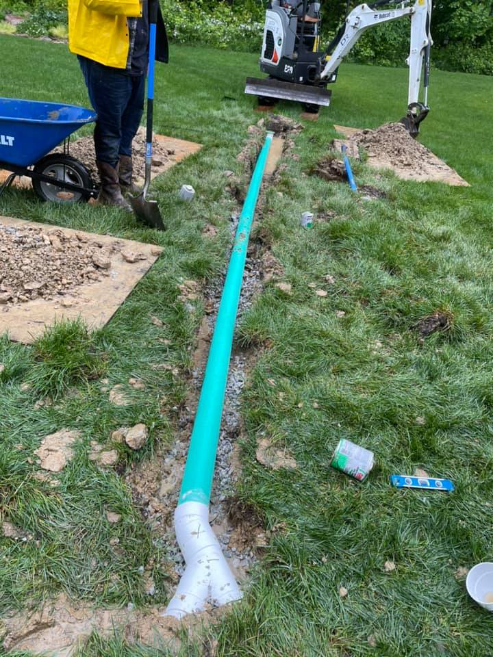 A bright blue pipe is being installed in a shallow trench in a grassy yard, with a worker holding a shovel and a mini excavator nearby.