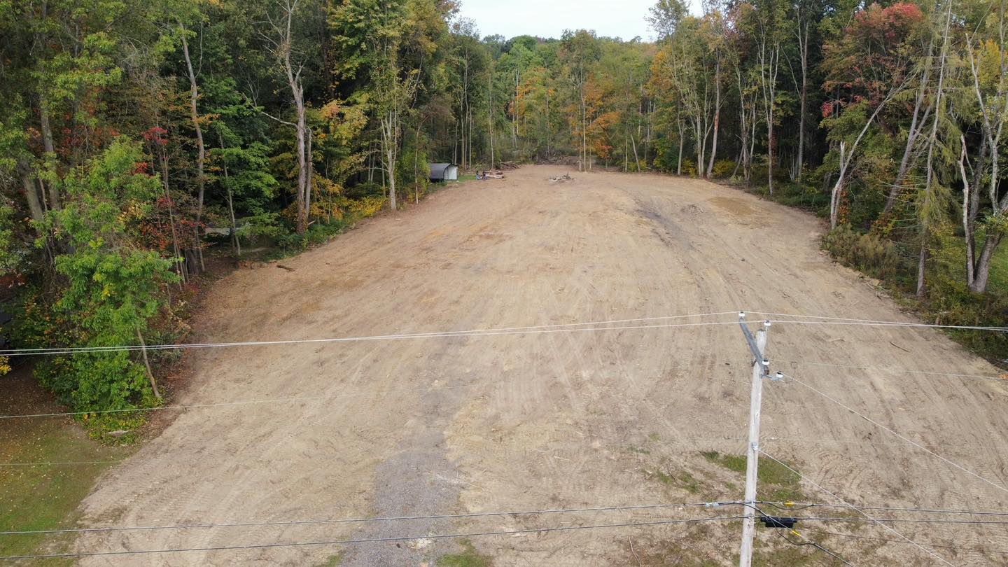 A cleared, dirt-covered lot surrounded by tall trees, with utility poles and wires in the foreground and a small structure in the background.