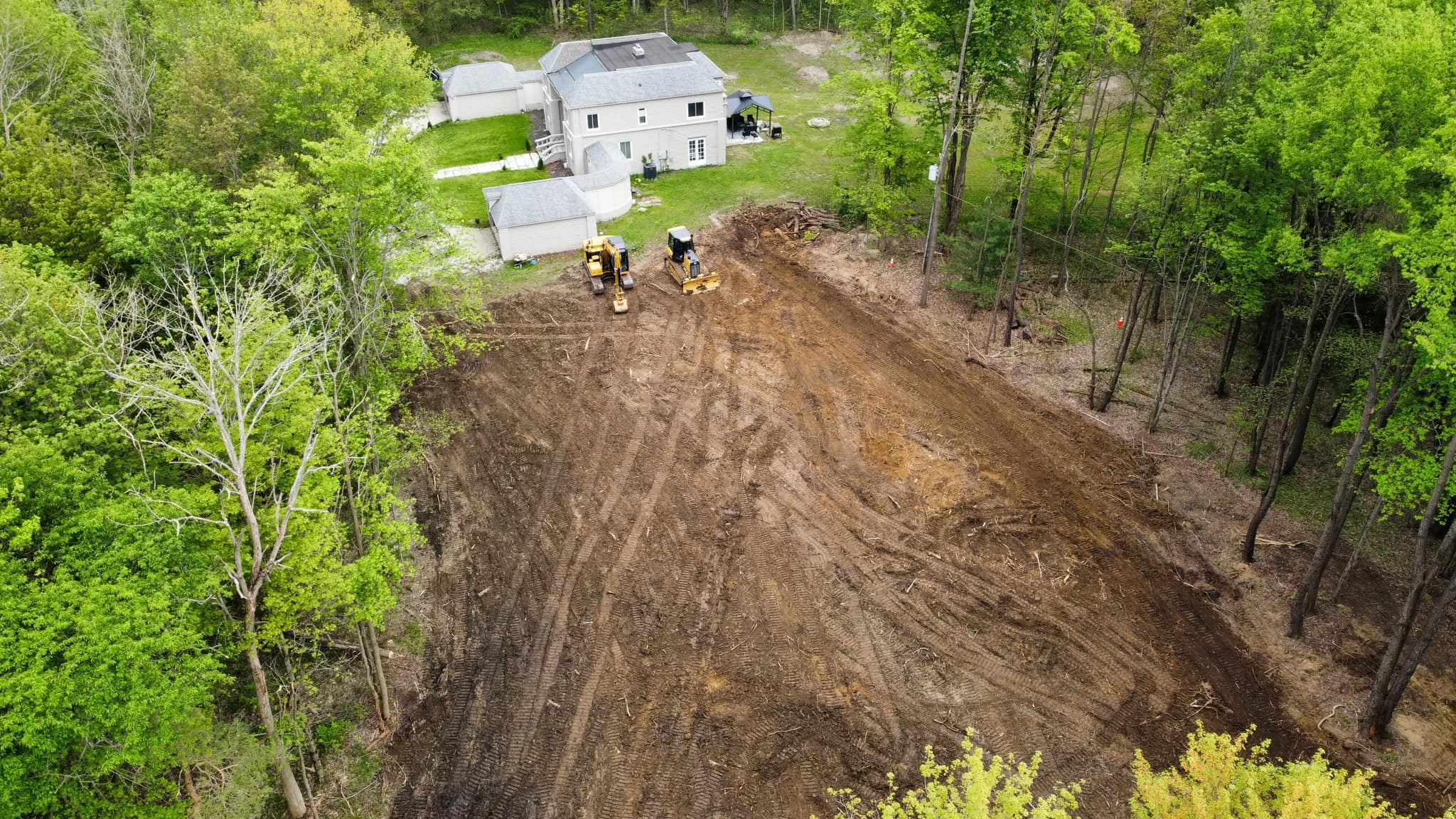 Aerial view of a cleared dirt area next to a house and garage, with an excavating contractor’s equipment and workers present, all surrounded by green trees.