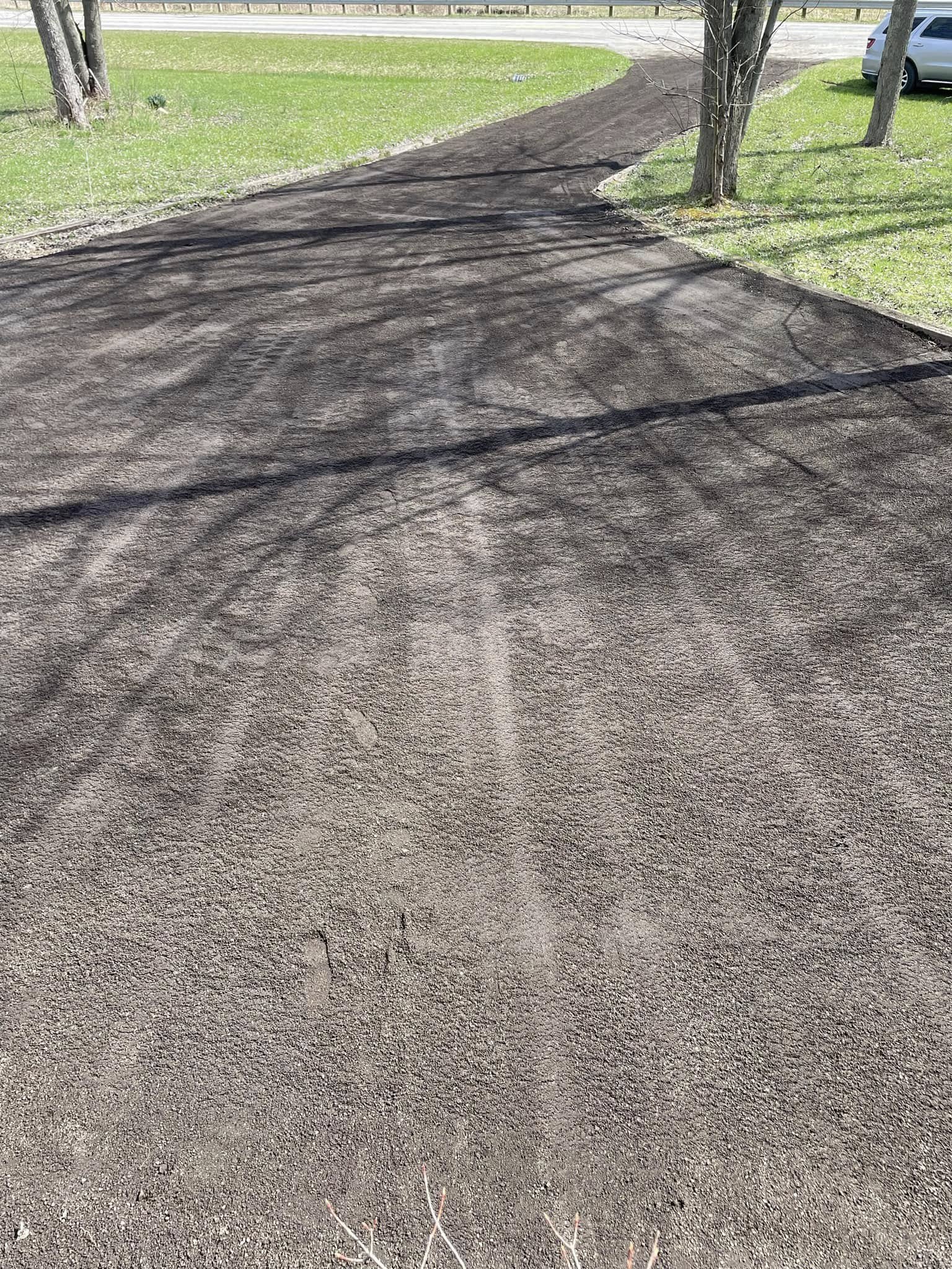 A newly resurfaced dirt driveway curves through a grassy area with trees and a parked car visible to the right.