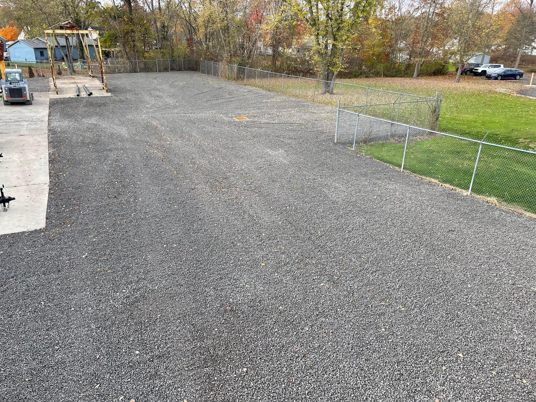 A large, fenced area covered with gravel next to a grassy yard, bordered by trees with autumn foliage and some playground equipment on the left.