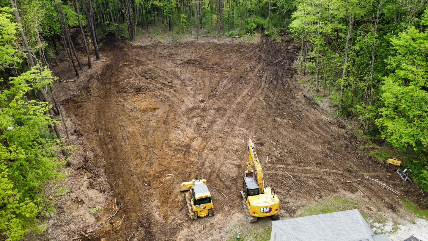 Aerial view of a cleared, leveled area in a forest with two yellow construction vehicles working near the edge of the site.