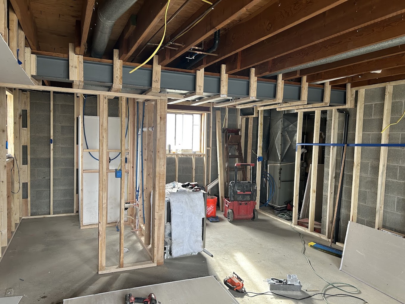 Room under construction with exposed wooden framing, unfinished walls, visible ceiling beams, tools, and construction materials scattered on the floor. Natural light enters through a window.