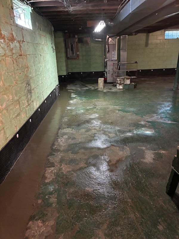 A flooded concrete basement floor with a professional waterproofing installation, featuring a perimeter drainage system along the green block wall.