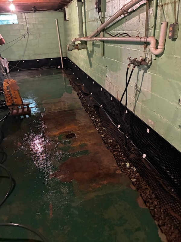A wet basement floor along a green concrete wall during a waterproofing installation, featuring a drainage channel and pipes.