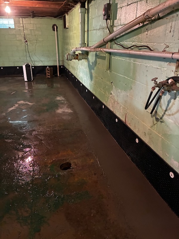 A basement interior featuring a waterproofing installation along the base of the mint-green cinder block wall, with a damp concrete floor and exposed overhead pipes.
