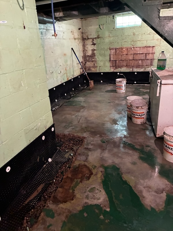 Basement interior with exposed block walls, a black drainage membrane installed for waterproofing installation along the foundation, and several buckets on a damp concrete floor.